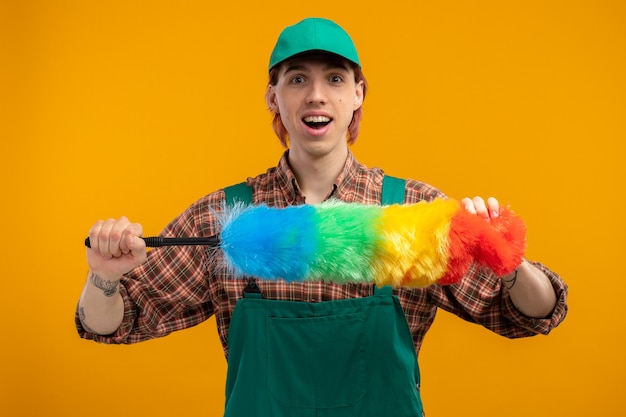 Hombre de limpieza joven feliz y positivo en mono de camisa a cuadros y gorra sosteniendo plumero colorido mirando sonriendo alegremente