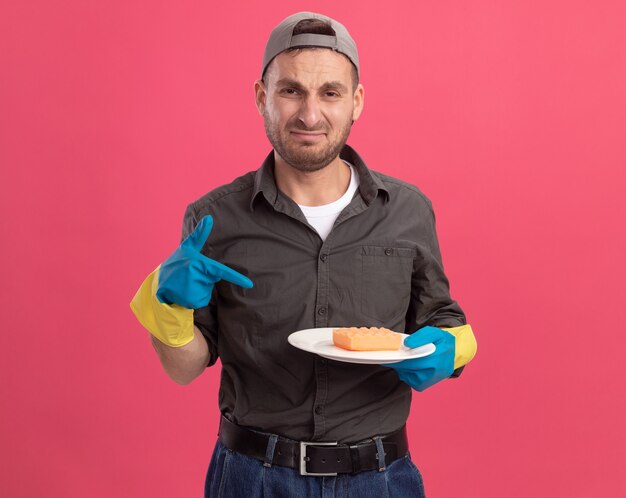 Hombre de limpieza joven molesto con ropa casual y gorra en guantes de goma que sostiene el plato y una esponja apuntando con el dedo índice al plato llorando frunciendo los labios sobre la pared rosa