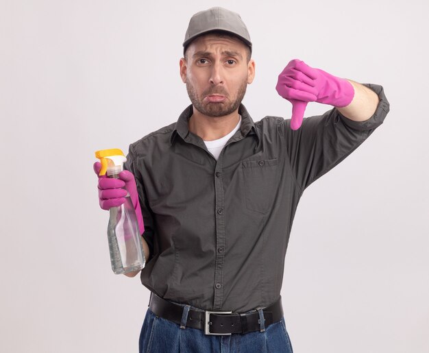 Foto gratuita hombre de limpieza joven molesto con ropa casual y gorra en guantes de goma sosteniendo una botella de spray mirando con expresión triste mostrando los pulgares hacia abajo de pie sobre la pared blanca