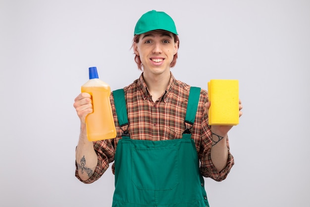 Hombre de limpieza joven en mono de camisa a cuadros y gorra con esponja y botella con productos de limpieza mirando con sonrisa en la cara