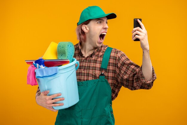 Hombre de limpieza joven en mono de camisa a cuadros y gorra sosteniendo un cubo con herramientas de limpieza gritando con expresión agresiva mientras habla por teléfono móvil de pie en naranja