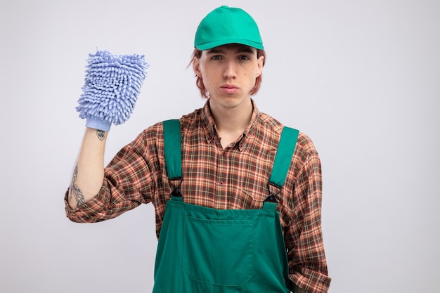 Hombre de limpieza joven en mono de camisa a cuadros y gorra sosteniendo plumero con cara seria listo para la limpieza de pie sobre la pared blanca