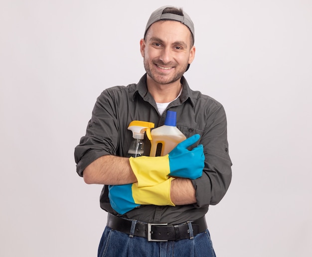Hombre de limpieza joven con ropa casual y gorra en guantes de goma con botella de spray y suministros de limpieza mirando sonriendo alegremente de pie sobre la pared blanca