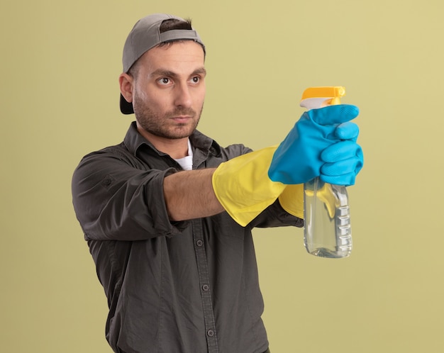 Foto gratuita hombre de limpieza joven con ropa casual y gorra en guantes de goma con el objetivo de la botella de spray mirando a un lado con la cara seria de pie sobre la pared verde