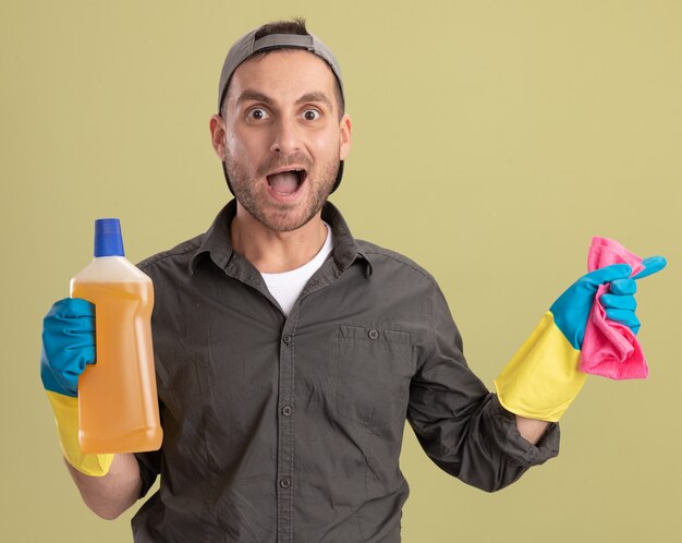 Hombre de limpieza joven con ropa casual y gorra en guantes de goma sosteniendo una botella con productos de limpieza y un trapo que parece feliz y emocionado de pie sobre la pared verde