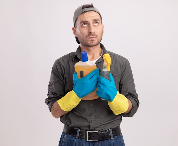 Hombre de limpieza joven con ropa casual y gorra en guantes de goma sosteniendo una botella de spray y artículos de limpieza mirando hacia arriba desconcertado parado sobre la pared blanca