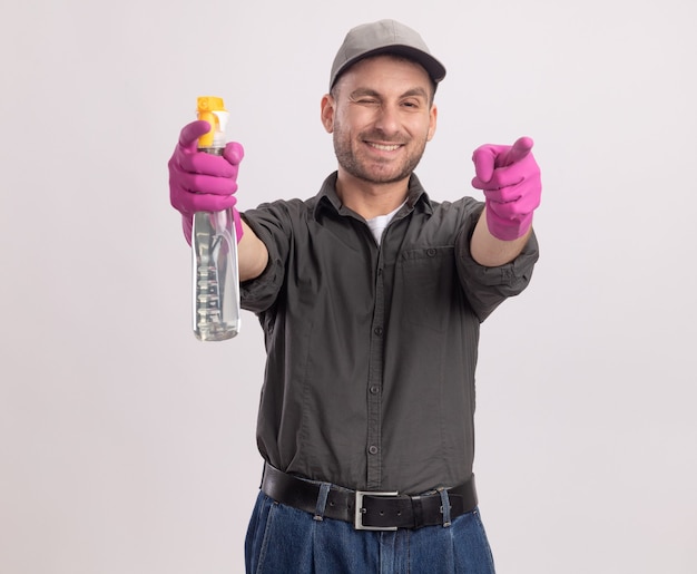 Hombre de limpieza joven con ropa casual y gorra en guantes de goma sosteniendo una botella de spray sonriendo y guiñando un ojo apuntando con los dedos índices de pie sobre la pared blanca