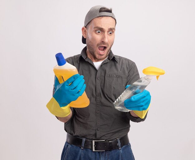 Hombre de limpieza joven con ropa casual y gorra en guantes de goma sosteniendo una botella de spray y suministros de limpieza asombrado y sorprendido de pie sobre la pared blanca