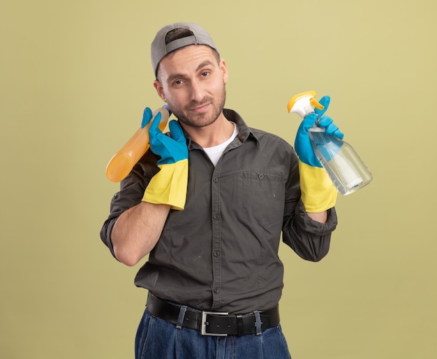 Foto gratuita hombre de limpieza joven con ropa casual y gorra en guantes de goma sosteniendo una botella de spray y suministros de limpieza mirando sonriente de pie confiado sobre la pared verde