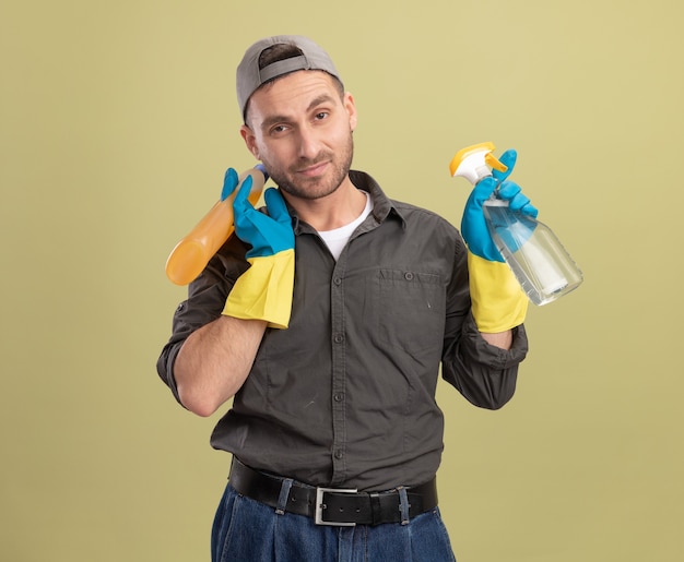 Hombre de limpieza joven con ropa casual y gorra en guantes de goma sosteniendo una botella de spray y suministros de limpieza mirando sonriente de pie confiado sobre la pared verde
