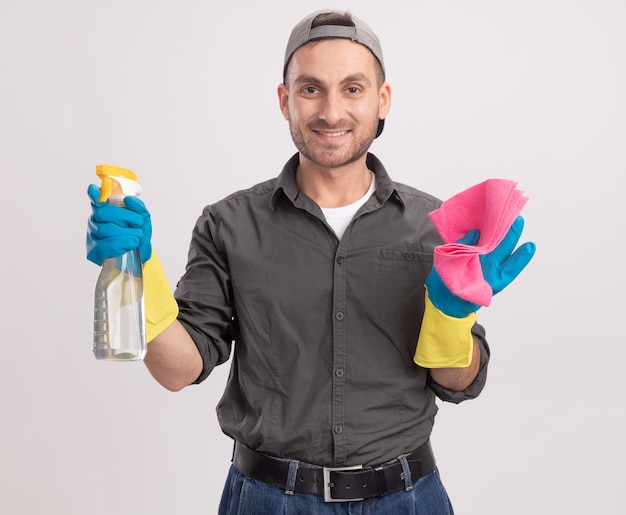 Hombre de limpieza joven con ropa casual y gorra en guantes de goma sosteniendo una botella de spray y un trapo con una sonrisa feliz y positiva de pie sobre la pared blanca