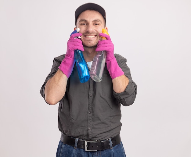 Foto gratuita hombre de limpieza joven con ropa casual y gorra en guantes de goma sosteniendo botellas de spray mirando sonriendo alegremente de pie sobre la pared blanca