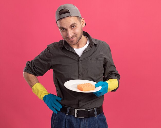 Hombre de limpieza joven con ropa casual y gorra en guantes de goma sosteniendo la placa y la esponja mirando sonriendo astutamente de pie sobre la pared rosa