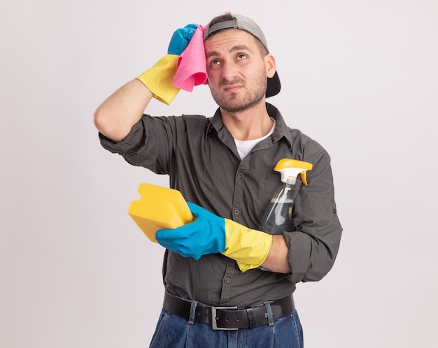 Hombre de limpieza joven con ropa casual y gorra en guantes de goma sosteniendo spray de limpieza y una esponja con un trapo en su hombro mirando hacia arriba desconcertado parado sobre la pared naranja