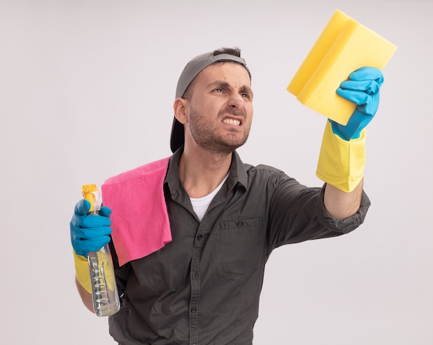 Hombre de limpieza joven con ropa casual y gorra en guantes de goma sosteniendo spray de limpieza y una esponja con un trapo en su hombro mirando la esponja con expresión molesta de pie sobre la pared naranja