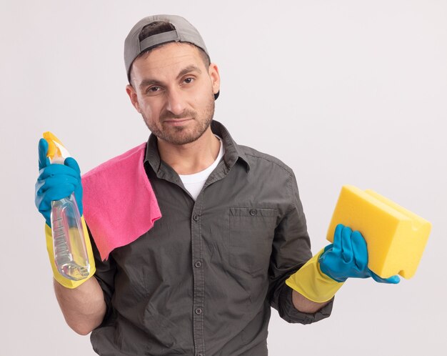 Hombre de limpieza joven con ropa casual y gorra en guantes de goma con spray de limpieza y una esponja con un trapo en su hombro que parece estar disgustado de pie sobre una pared naranja