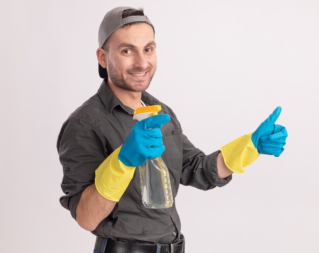 Hombre de limpieza joven con ropa casual y gorra en guantes de goma con spray de limpieza mirando sonriendo mostrando los pulgares para arriba sobre la pared naranja
