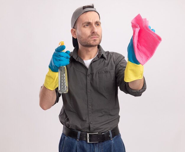 Hombre de limpieza joven con ropa casual y gorra en guantes de goma con spray de limpieza y un trapo mirando a un lado de cerca con la cara seria de pie sobre la pared blanca
