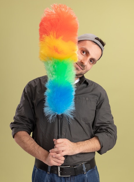 Foto gratuita hombre de limpieza joven con ropa casual y gorra sosteniendo un plumero colorido que parece feliz y alegre de pie sobre la pared verde