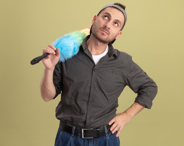 Hombre de limpieza joven vistiendo ropa casual y gorra sosteniendo plumero colorido mirando hacia arriba desconcertado de pie sobre la pared verde