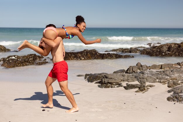 Hombre llevando a mujer sobre los hombros en la playa bajo el sol