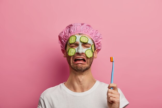 Hombre llorando insatisfecho sostiene cepillo de dientes, aplica mascarilla facial de arcilla con pepinos, cansado de tratamientos de belleza