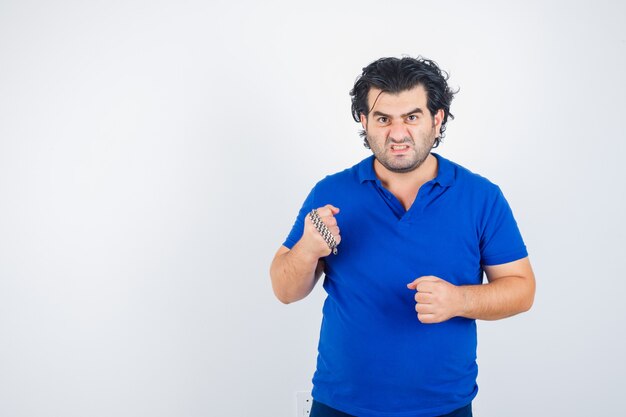 Hombre maduro amenazando con cadena envuelta por puño, apretando los dientes en camiseta azul y luciendo agresivo. vista frontal.