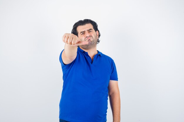 Hombre maduro apuntando a un lado con el pulgar en camiseta azul, mirando decepcionado, vista frontal.