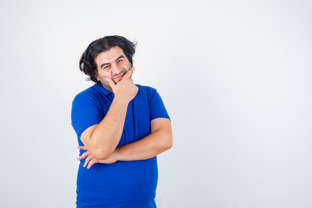 Hombre maduro, con la barbilla apoyada en la palma, pensando en algo en camiseta azul, jeans y mirando pensativo, vista frontal.