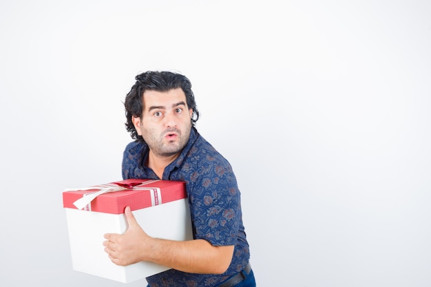 Hombre maduro con caja de regalo en camisa y mirando perplejo, vista frontal.