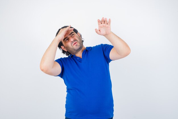 Hombre maduro en camiseta azul, jeans mirando lejos con la mano sobre la cabeza, estirando la mano y mirando enfocado, vista frontal.