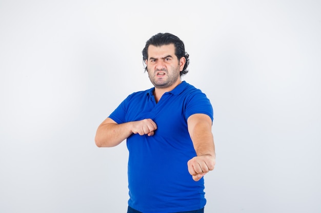 Hombre maduro en camiseta azul, jeans de pie en pose de lucha y mirando enojado, vista frontal.