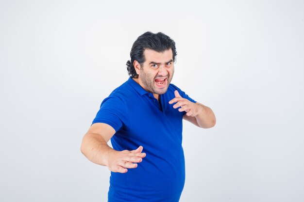 Hombre maduro en camiseta azul, jeans de pie en pose de lucha y mirando enojado, vista frontal.