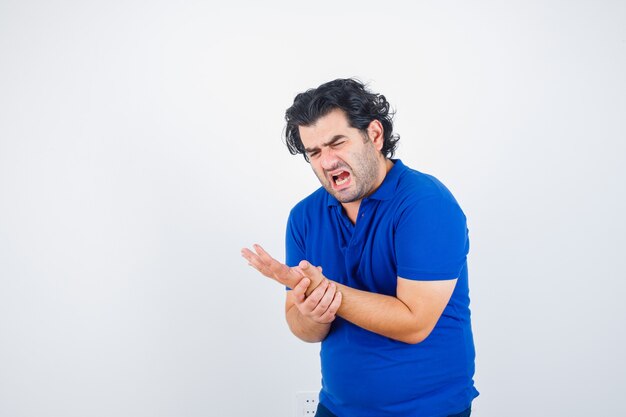 Hombre maduro en camiseta azul sosteniendo su muñeca dolorosa y mirando angustiado, vista frontal.