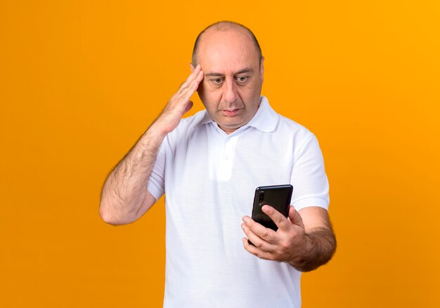 Hombre maduro casual confundido sosteniendo y mirando el teléfono poniendo la mano en el templo aislado sobre fondo amarillo