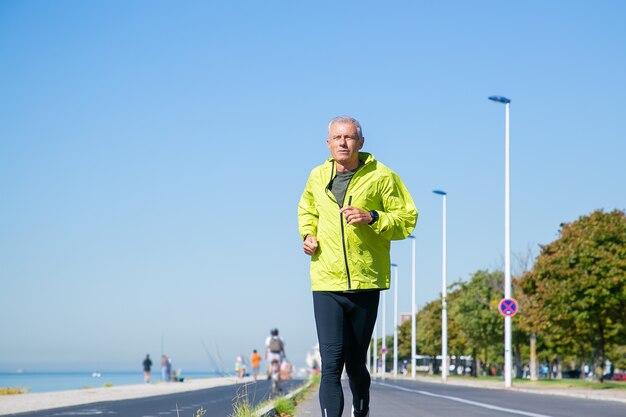Hombre maduro centrado en chaqueta deportiva verde y medias para correr a lo largo de la orilla del río en el exterior. Entrenamiento de corredor senior para maratón. Vista frontal. Concepto de actividad y edad