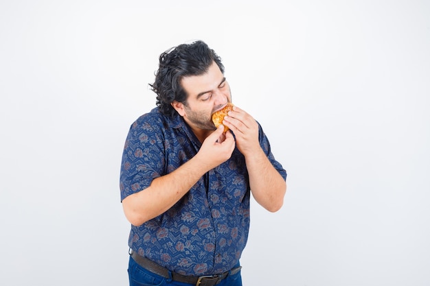 Hombre maduro comiendo producto de pastelería en camisa y mirando encantado, vista frontal.