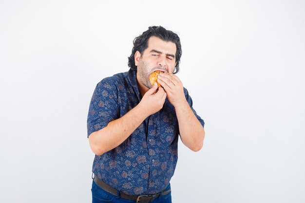 Hombre maduro comiendo producto de pastelería en camisa y mirando encantado, vista frontal.