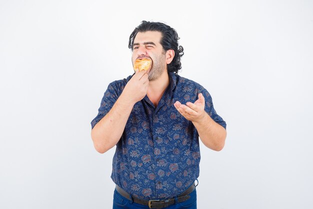 Hombre maduro comiendo productos de pastelería mientras estira la mano en gesto de interrogación en camisa y mirando encantado, vista frontal.
