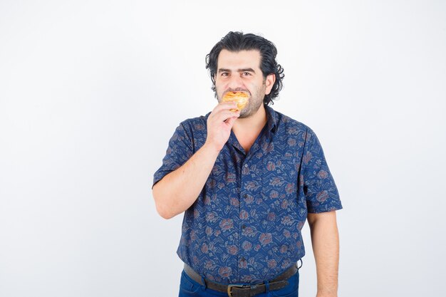 Hombre maduro comiendo productos de pastelería mientras mira a la cámara en camisa y mira encantada, vista frontal.