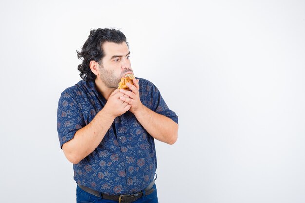 Hombre maduro comiendo productos de pastelería mientras mira a otro lado en camisa y parece desconcertado. vista frontal.