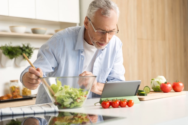 Hombre maduro concentrado cocinando ensalada usando tableta