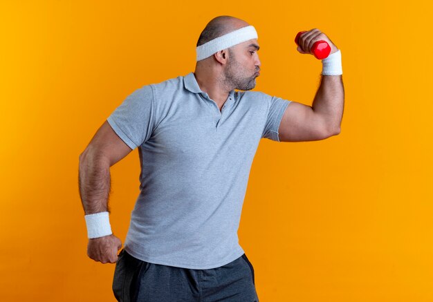 Hombre maduro deportivo en diadema levantando la mano con mancuernas mostrando bíceps mirando confiado de pie sobre la pared naranja