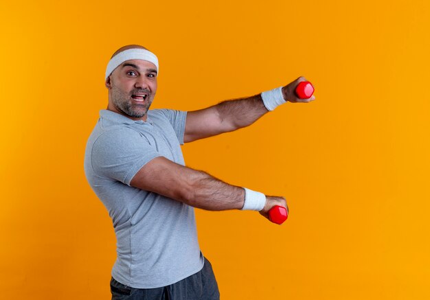 Hombre maduro deportivo en diadema trabajando con pesas mirando confiado sonriendo de pie sobre la pared naranja