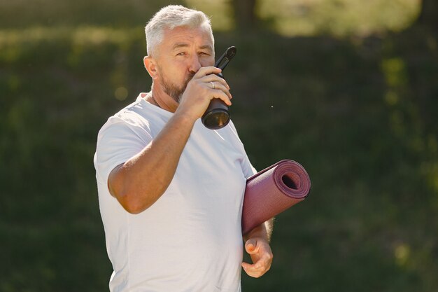 Hombre maduro entrenando en un parque de verano. Senior de pie con una estera. Anciano en ropa deportiva.