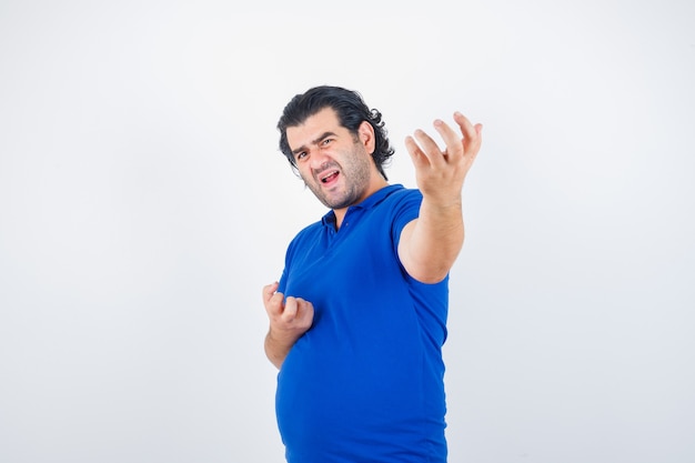 Hombre maduro estirando la mano como sosteniendo algo imaginario en camiseta azul, jeans y mirando enojado. vista frontal.