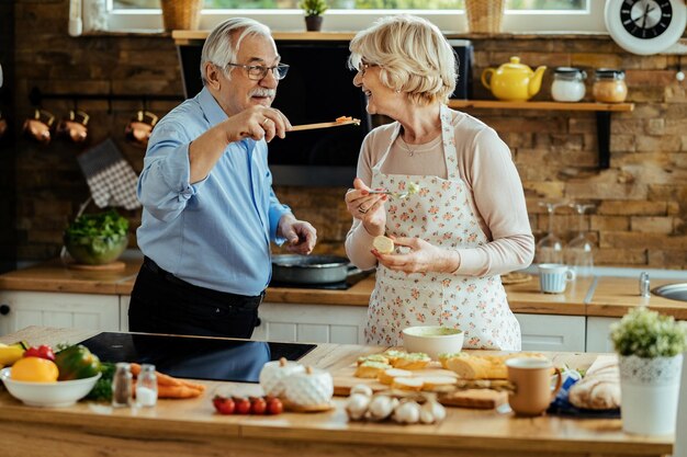 Hombre maduro feliz dando a su esposa para probar la comida que está cocinando en la cocina