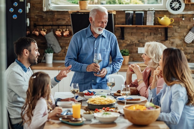 Hombre maduro feliz proponiendo un brindis y hablando con su familia durante el almuerzo en el comedor.