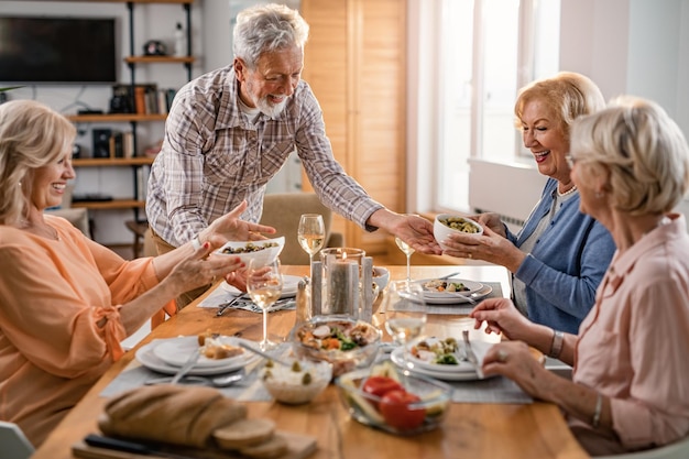 Hombre maduro feliz sirviendo comida mientras almuerza con amigas en casa