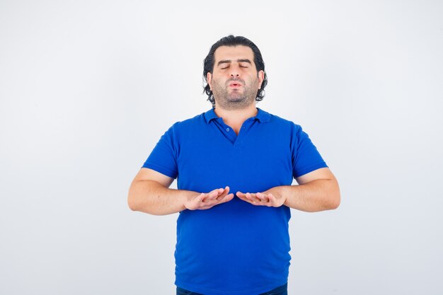 Hombre maduro meditando, manteniendo los ojos cerrados en camiseta azul, jeans y mirando tranquilo, vista frontal.